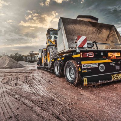 DOLL low-loader on a muddy construction site between piles of earth, loaded with a construction site vehicle