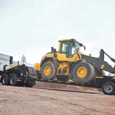 DOLL low-bed semi-trailer from the side, loaded with construction site vehicle, on sandy ground