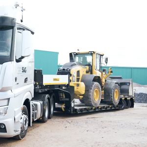 Un camion avec remorque surbaissée de DOLL, sur lequel est chargé un véhicule de chantier, prend un virage