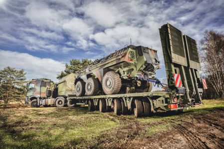 Militärischer Schwerlasttransporter mit getarntem gepanzerten Fahrzeug auf Tieflader in einer Waldlandschaft.