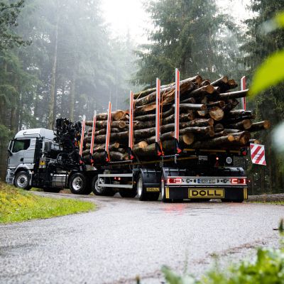 A silver logging truck with a loaded trailer drives on a rain-soaked road through a foggy forest.