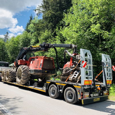 Heavy-duty tractor secured on a flatbed trailer for transportation through a forested road under a sunny sky.