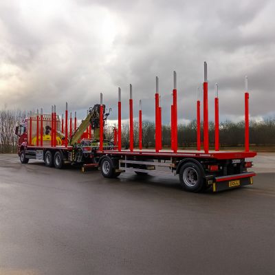 Red timber transport truck with a long log trailer, red stakes, and a loading crane on a wet road.