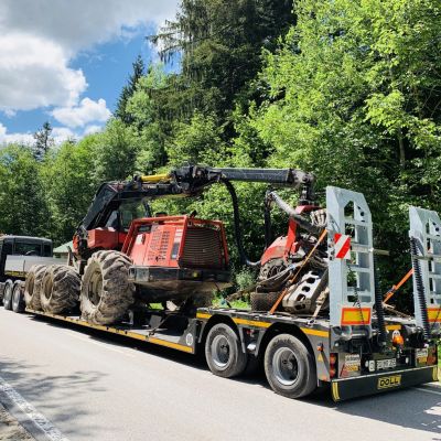 Heavy-duty tractor secured on a flatbed trailer for transportation through a forested road under a sunny sky.